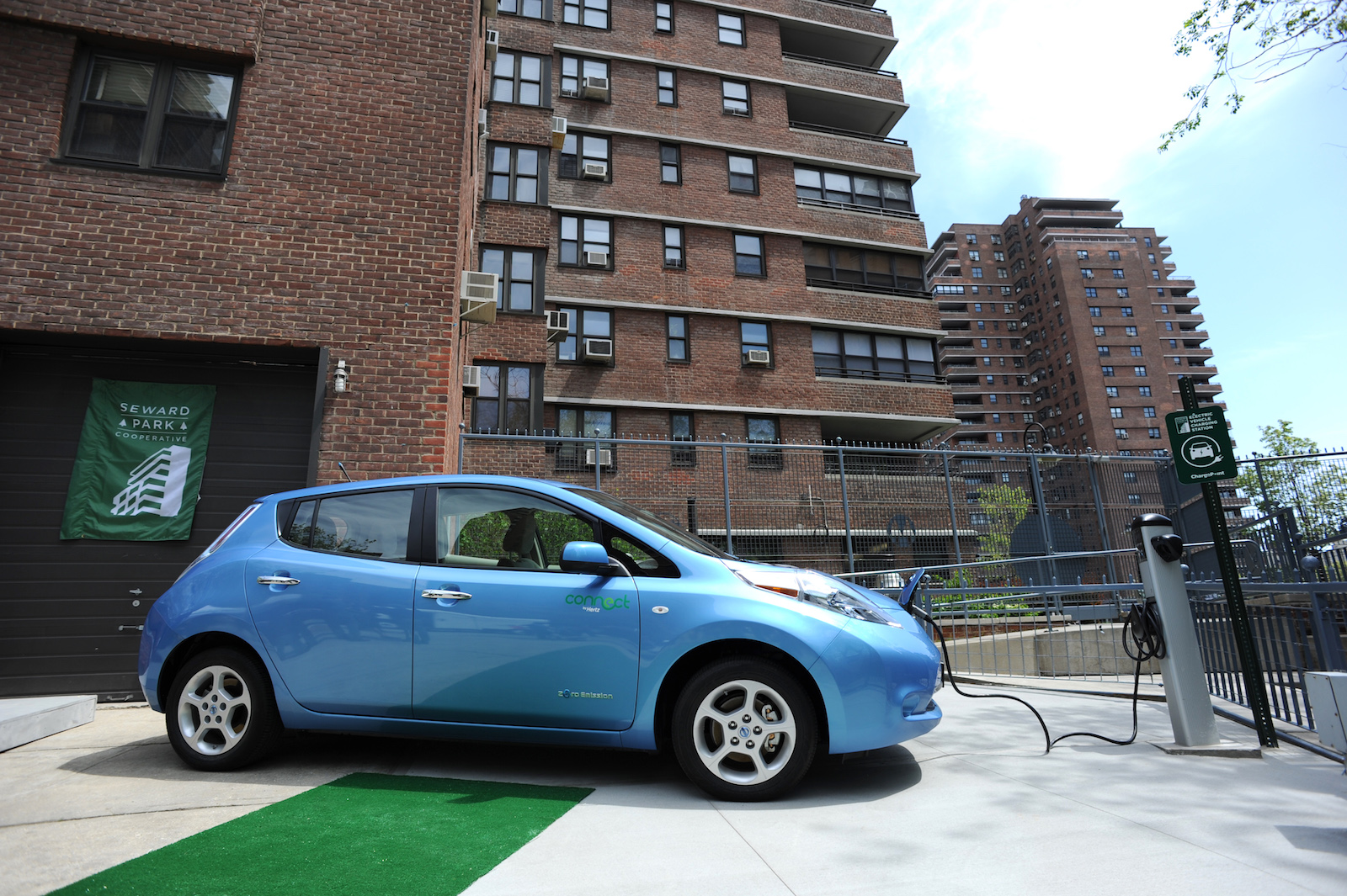 a blue Nissan Leaf is plugged into a charging station outside of a red brick apartment building with chain link fence surrounding it