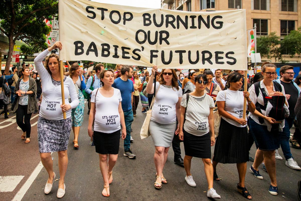 A line of pregnant women in white shirts carry a sign that says "stop burning our babies' futures."