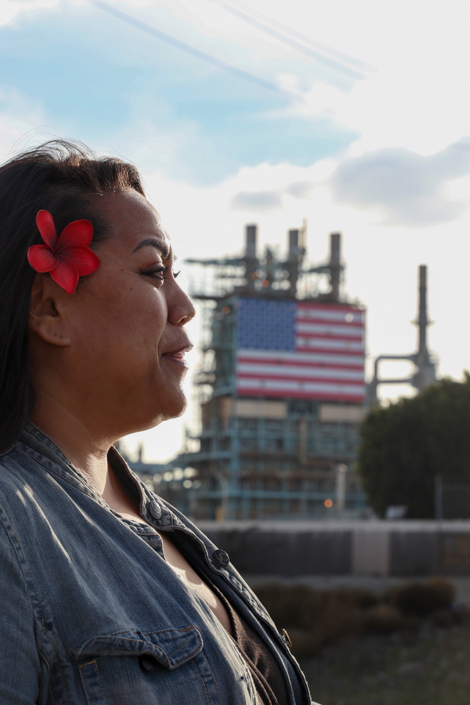 A woman with a flower in her hair looks to the right in profile with a large refinery with an American flag on it is seen in the background