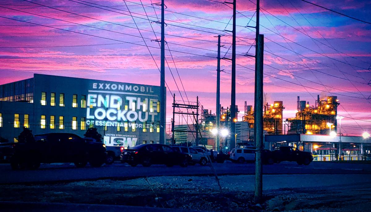 Oil refinery in texas lit up at night. The words "end the lockout" are projected onto the side of a building.