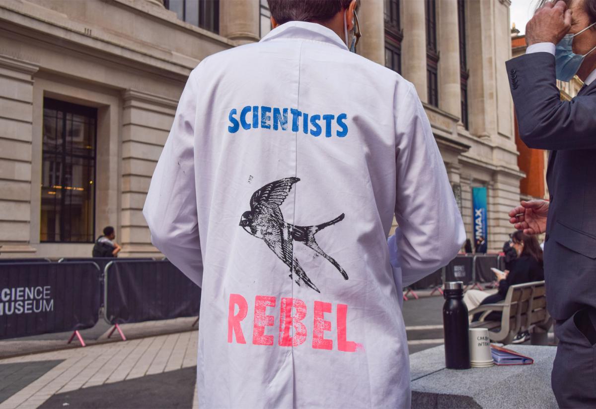 Scientist with back to camera; lab coat reads "Scientists Rebel" with an image of a swallow