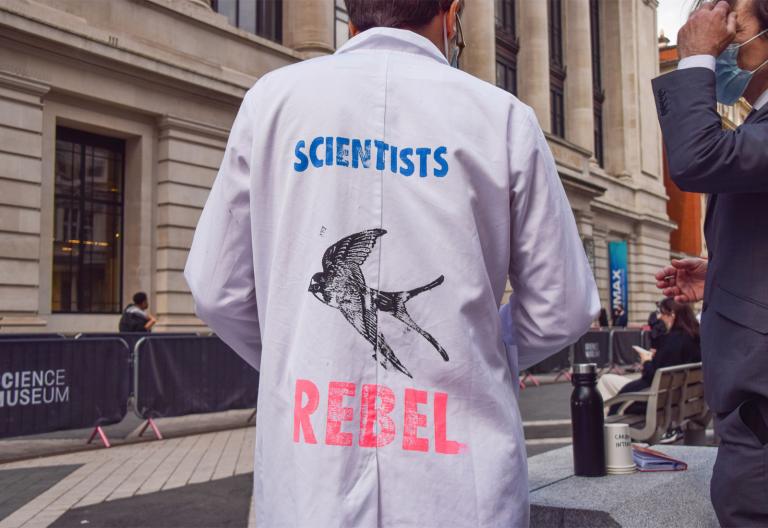 Scientist with back to camera; lab coat reads "Scientists Rebel" with an image of a swallow