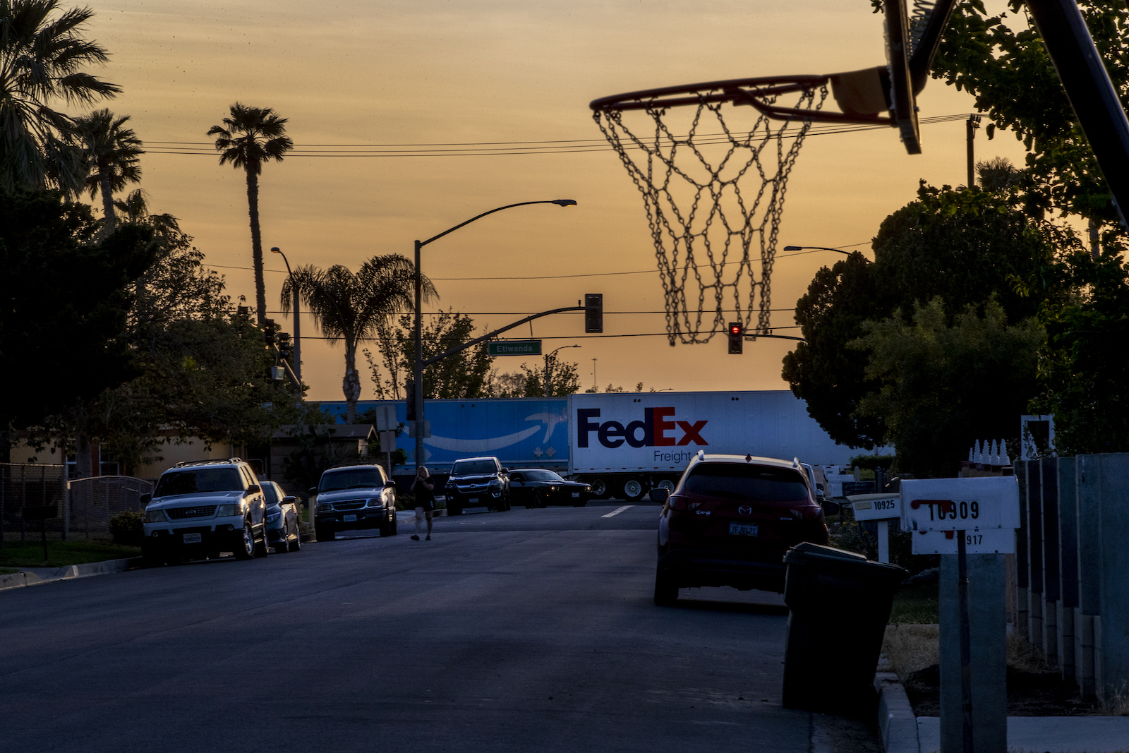 Two 18-wheeler trucks pass through a neighborhood below an orange sky with a basketball hoop in the foreground.