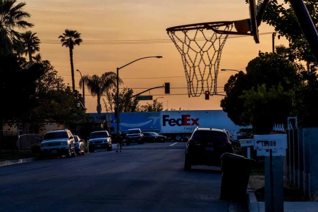 Two 18-wheeler trucks pass through a neighborhood below an orange sky with a basketball hoop in the foreground.