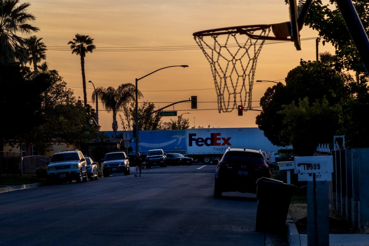 Two 18-wheeler trucks pass through a neighborhood below an orange sky with a basketball hoop in the foreground.