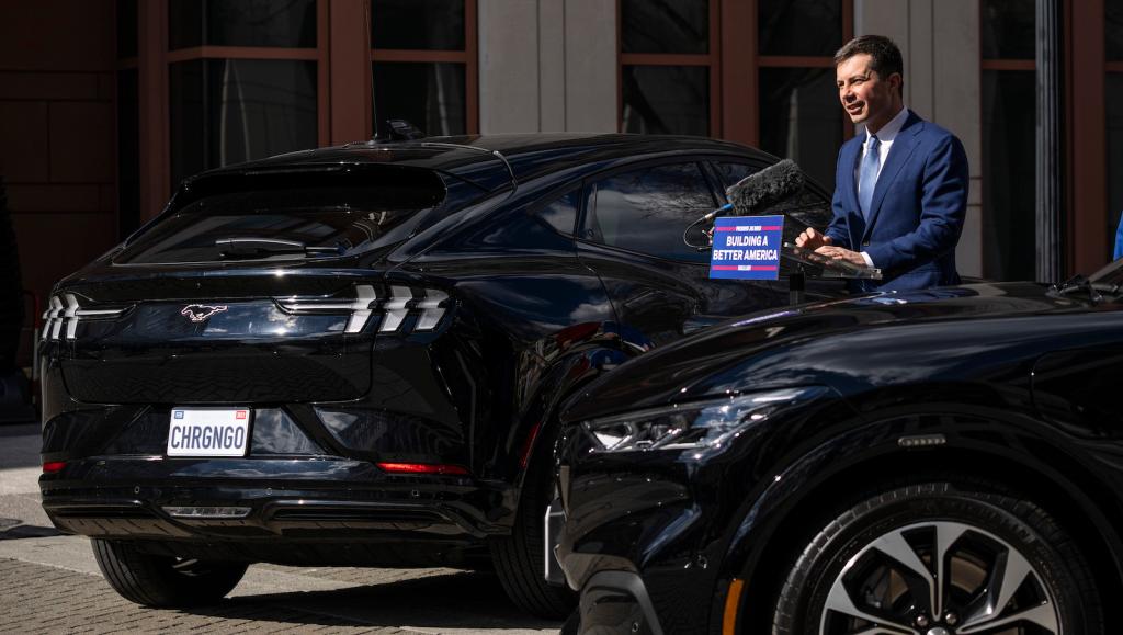 Pete Buttigieg stands between two black electric cars.