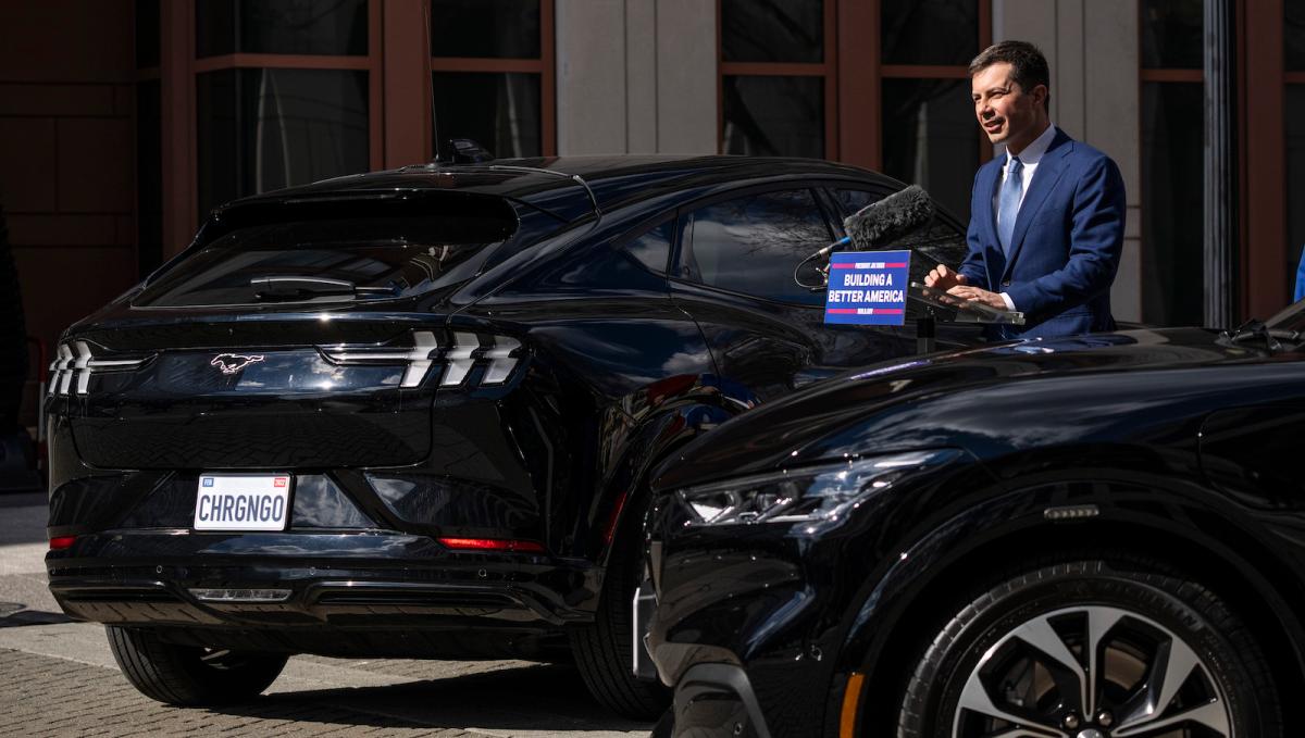 Pete Buttigieg stands between two black electric cars.