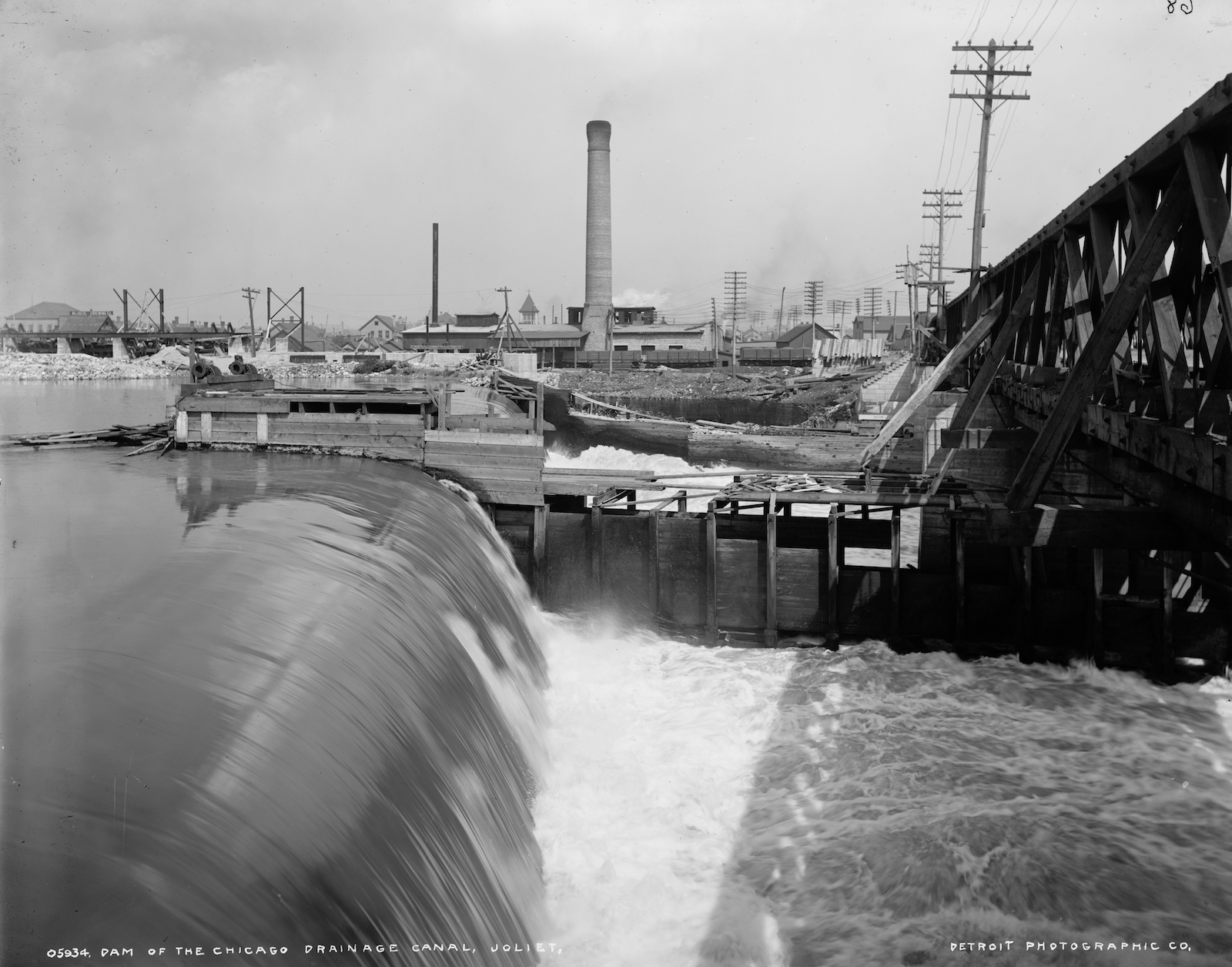 a black and white photo of a dam with a lot of water pouring from one level to another. In the background, a tall smokestack and industrial infrastructure
