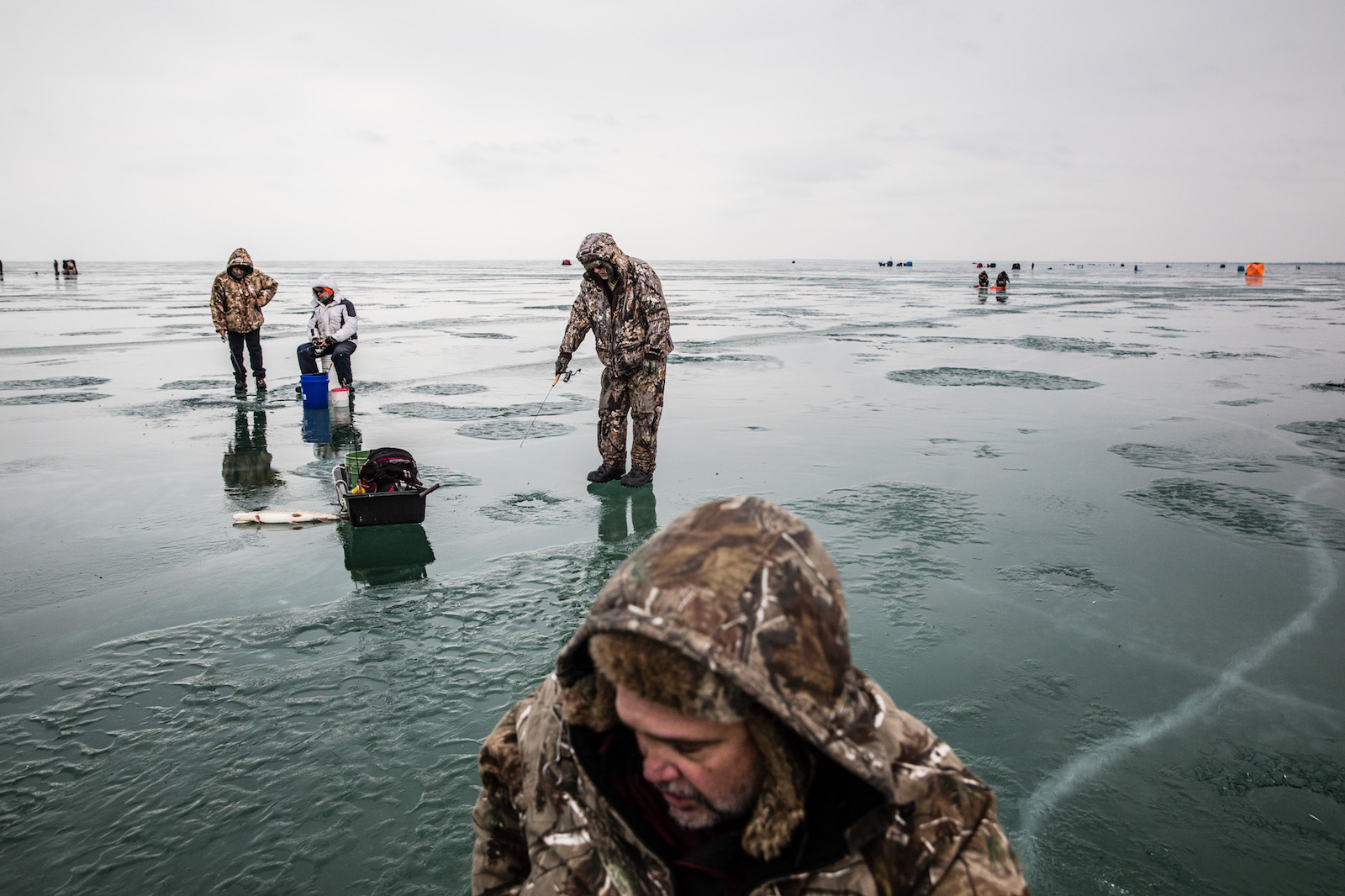 a group of people in camo gather on watery ice bending over buckets and holding fishing poles