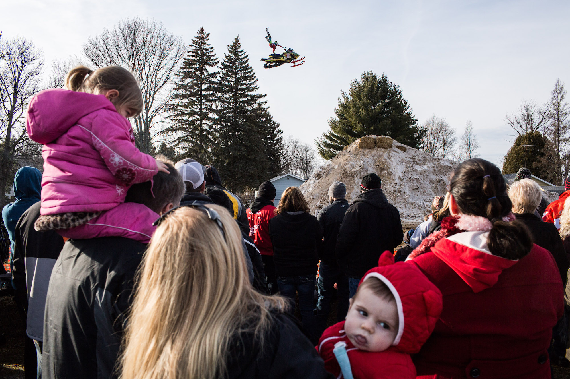 a snowmobile and rider fly through the air above a crowd of onlookers including small children in bright quilted jackets