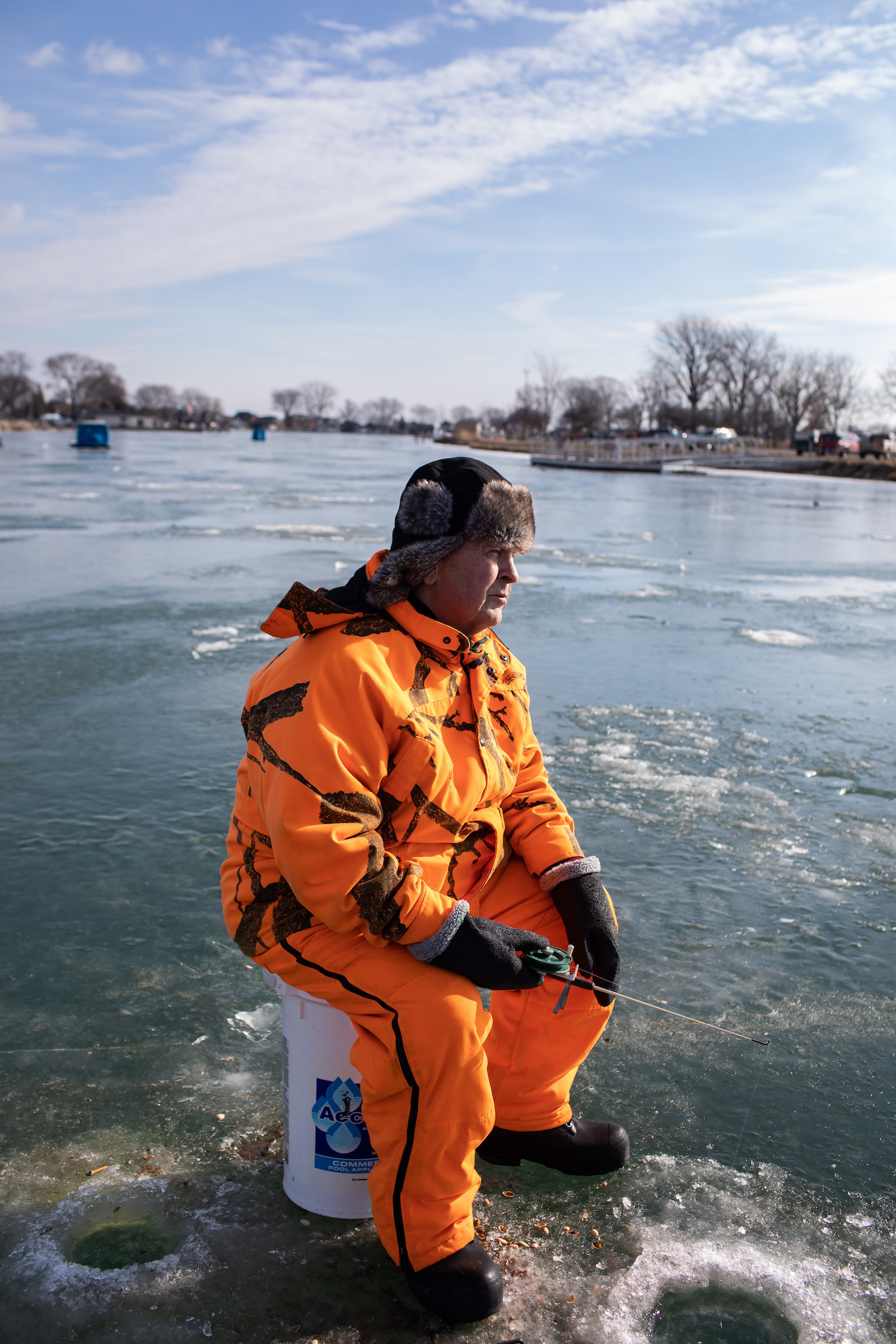 A man in an orange jumpsuit and furry hat sits on a bucket on the middle of a frozen lake