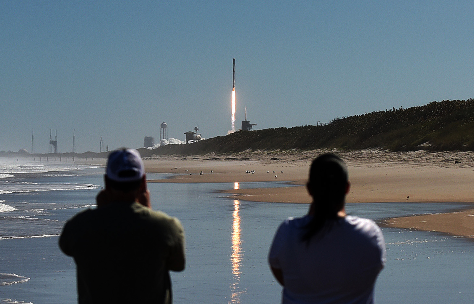 two people stand with their backs to the viewer watching a rocket take off over a seaside view
