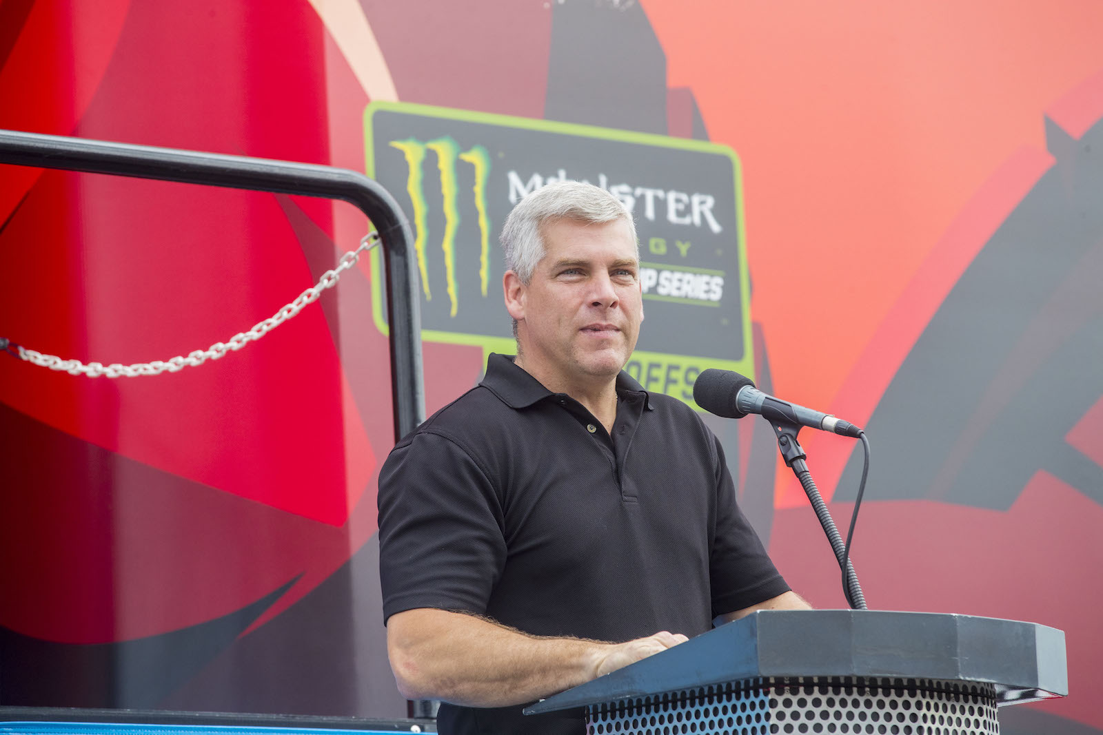 a man with white hair and a black polo shirt stands in front of a podium with a speaker