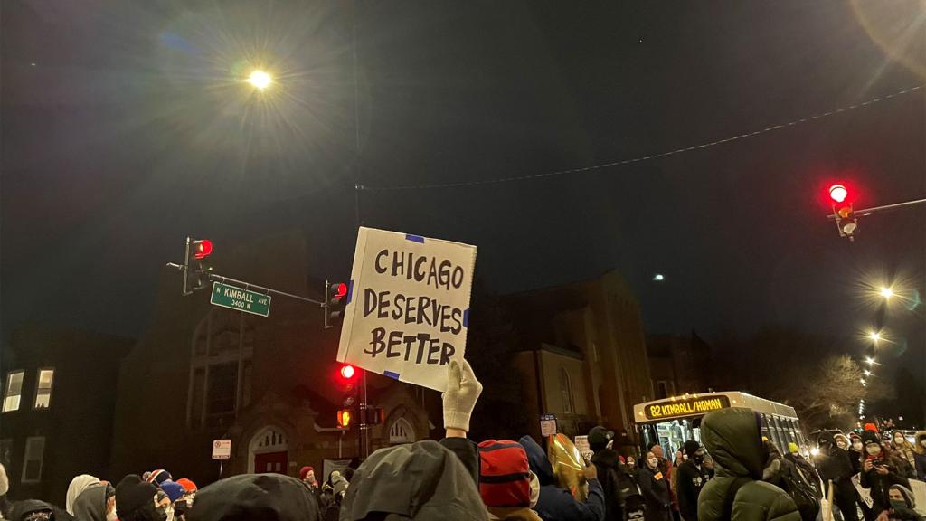Protesters in street with stoplights and bus in background; sign reading 