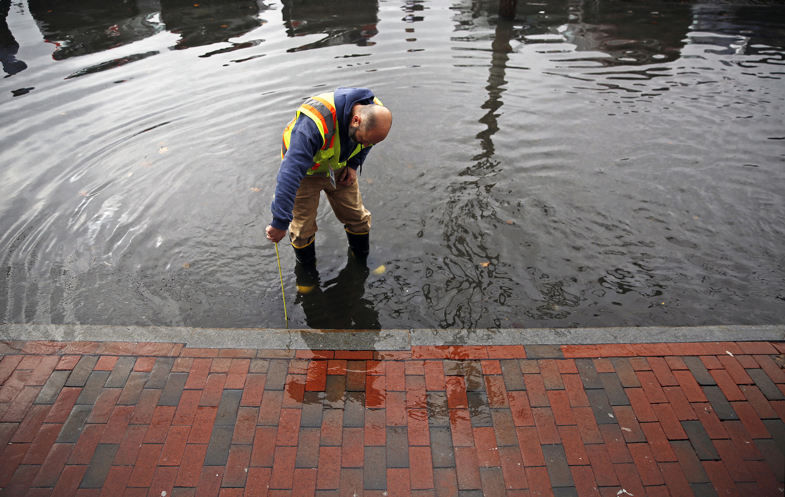 man in yellow construction vest bends over street flooded with water measuring depth with ruler. Bottom of the screen is red brick road