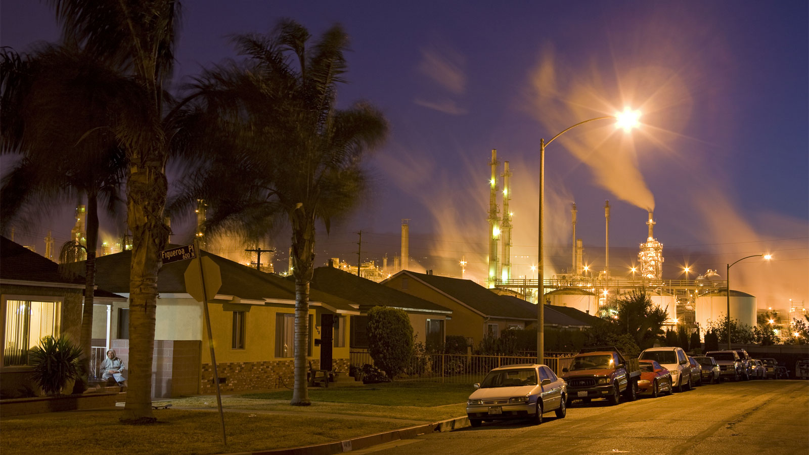 Residential street with palm trees, houses, and cars; oil refinery in near background