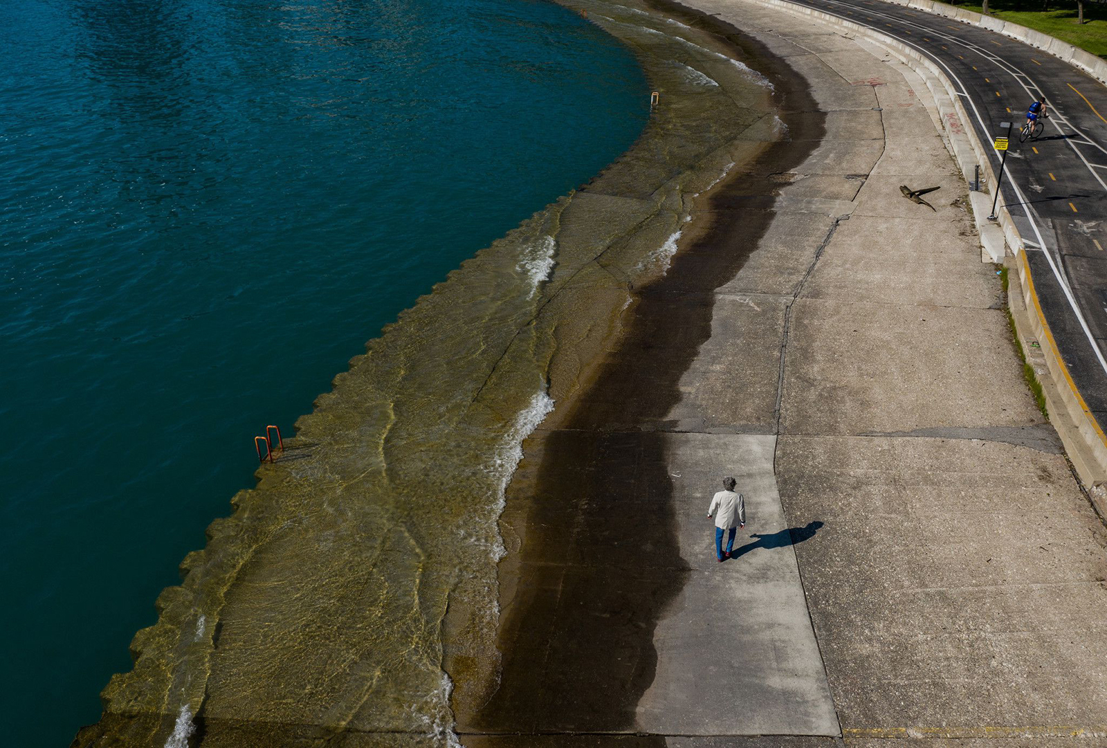 a person walks along the edge of a blue-green body of water on a road which is partially covered with water overflow