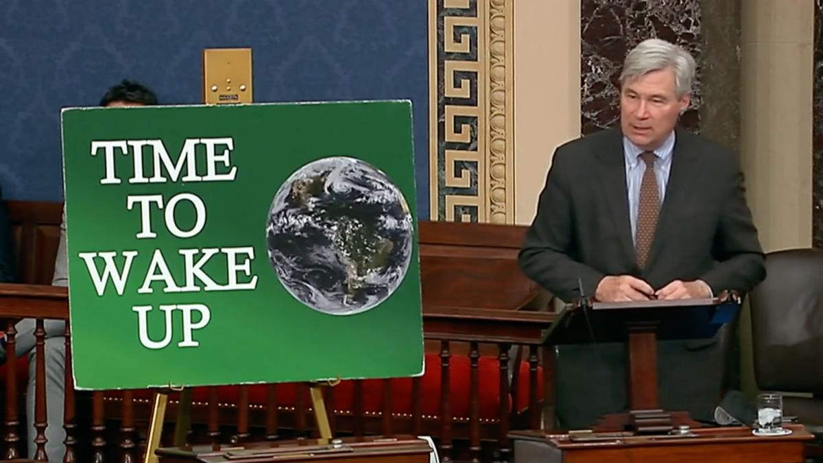 Senator Sheldon Whitehouse stands next to a green sign with a globe and the words "Time To Wake UP" on it.
