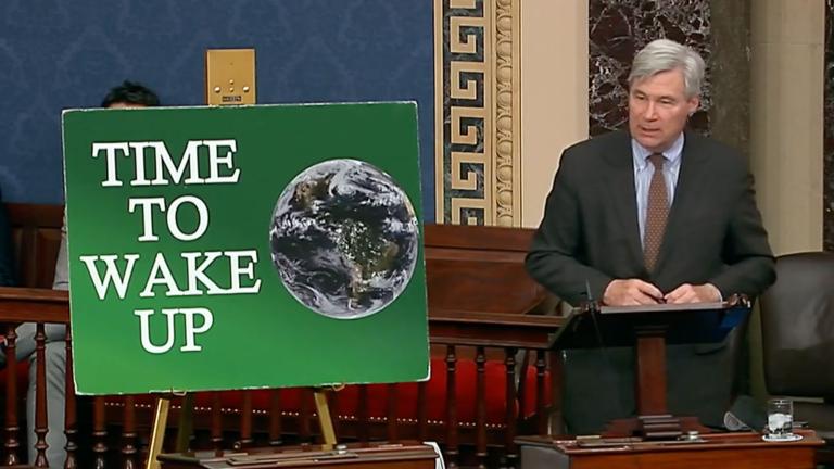 Senator Sheldon Whitehouse stands next to a green sign with a globe and the words "Time To Wake UP" on it.