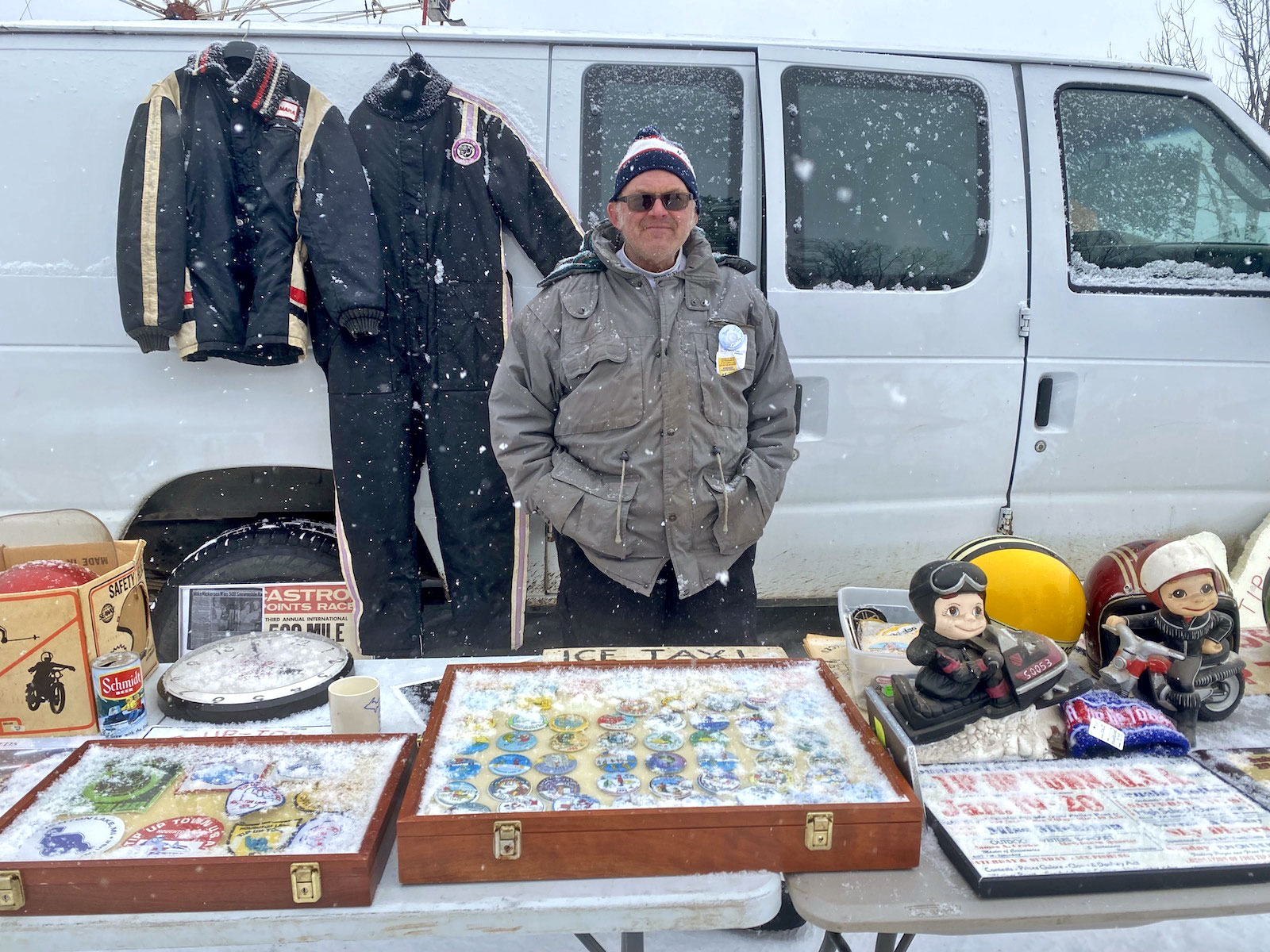 a man in a light gray-brown parka stands in front of a table laden with old buttons and plates and dolls. Behind him, a white van is parked with jackets hanging from the side.