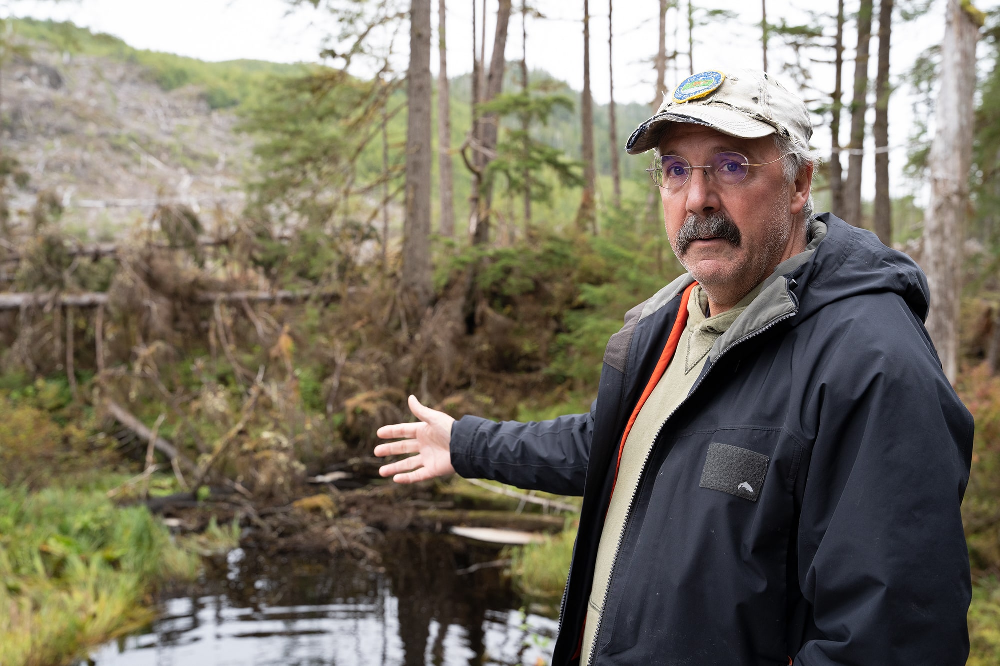 a man with glasses and a mustache wearing a black jacket gestures toward a river with felled trees