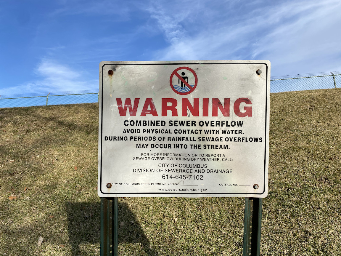 A sign reads: "WARNING COMBINED SEWER OVERFLOW AVOID PHYSICAL CONTACT WITH WATER. DURING PERIODS OF RAINFALL SEWAGE OVERFLOWS MAY OCCUR INTO THE STREAM."