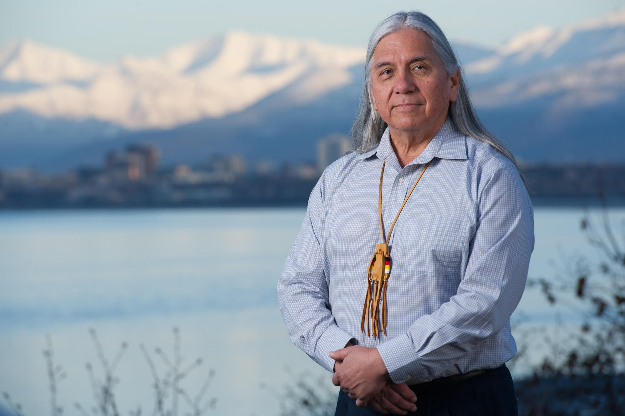 A man with long gray hair wearing a blue button-up shirt and leather necklace stands in front of a lake and snow-capped mountains