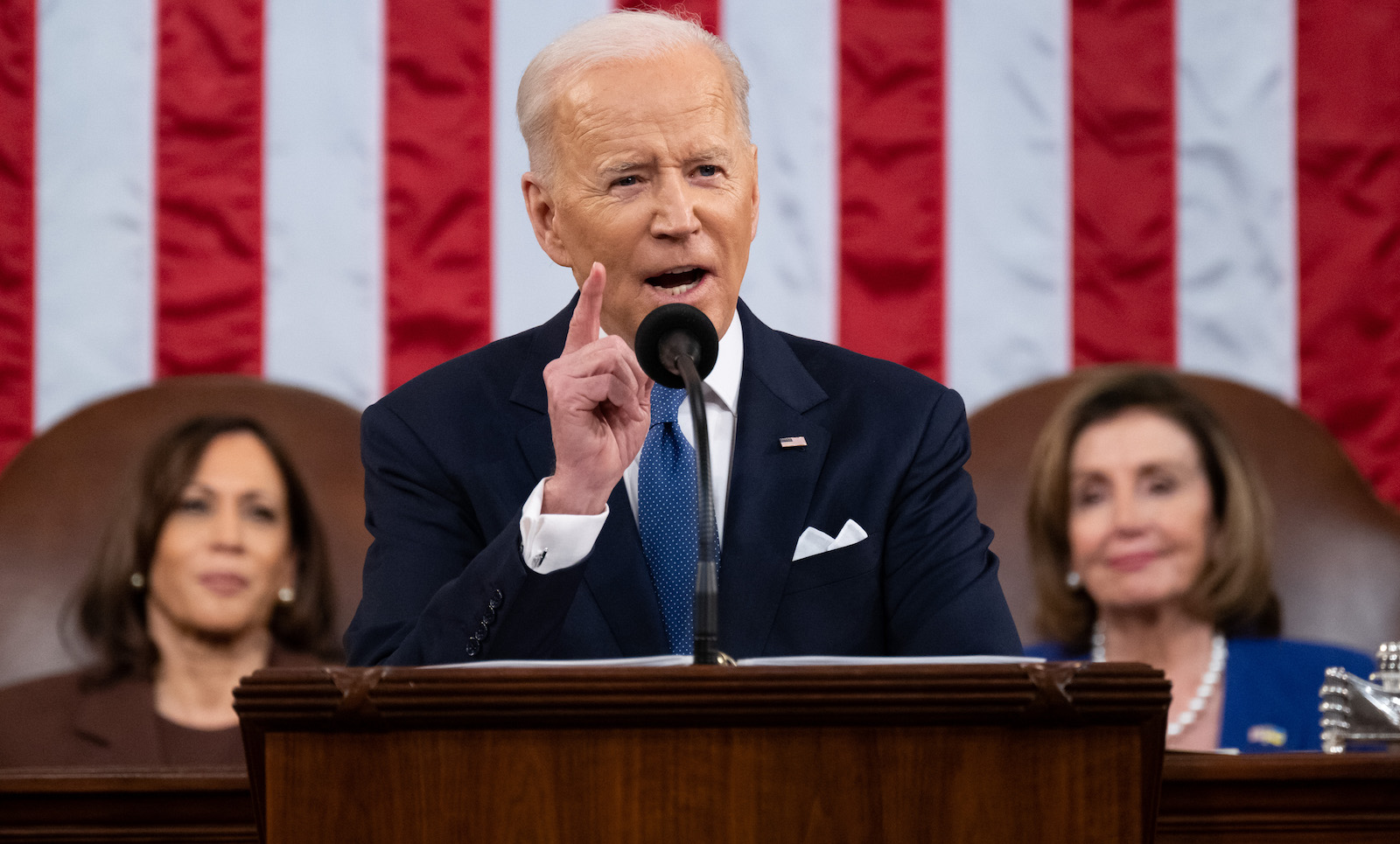 joe biden gestures with one hand in front of a podium during the 2022 state of the union speech while two women (kamala harris, left, and nancy pelosi, right) in suits sit behind him on either side