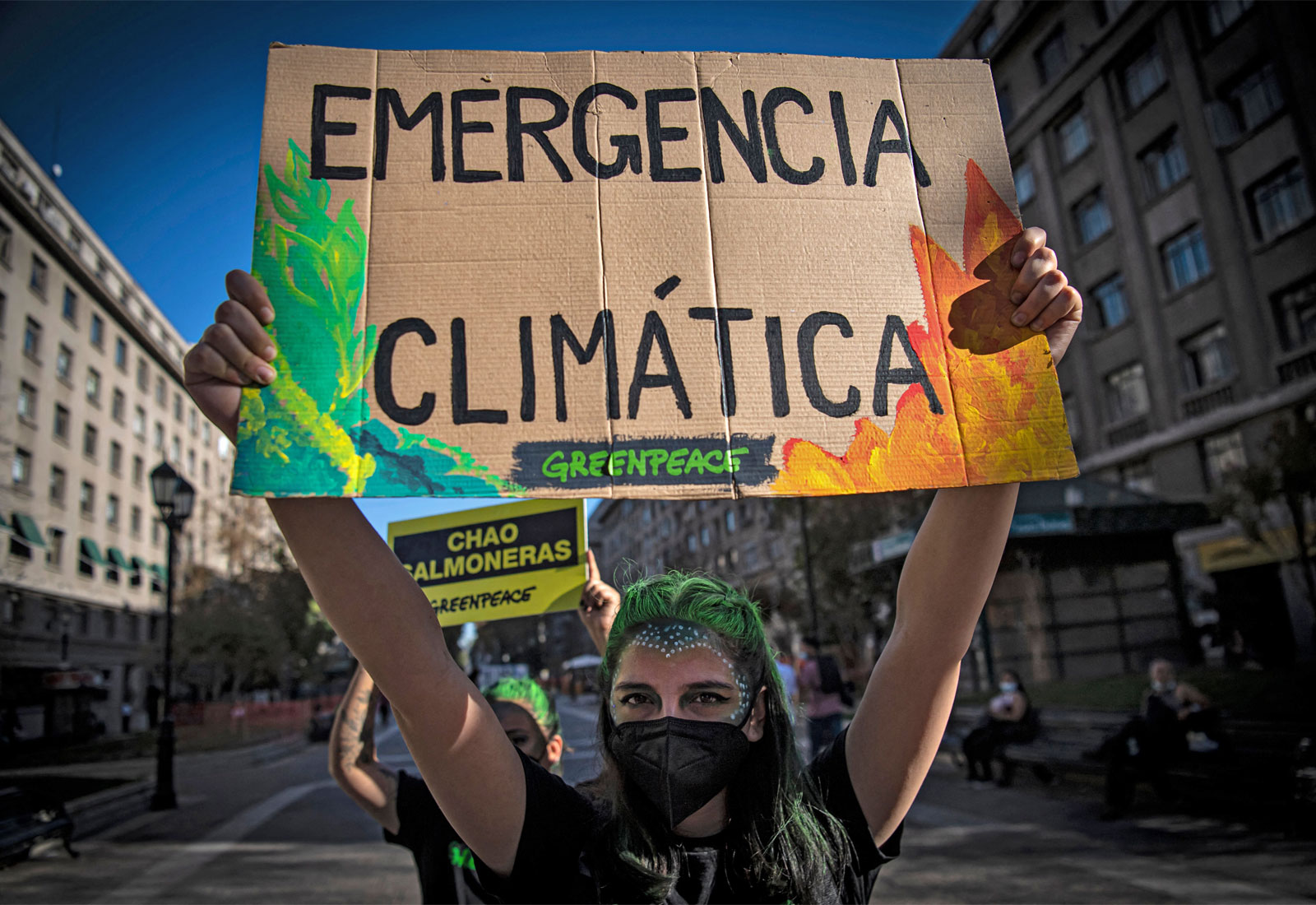 Woman with green hair and mask holding cardboard sign with leaves and flames that reads "emergencia climática"
