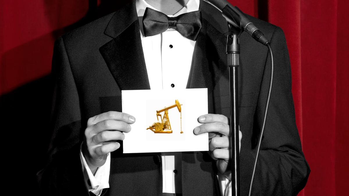 black and white photo of man in tux holding an envelope with a gold oil pump on it; red curtains in background