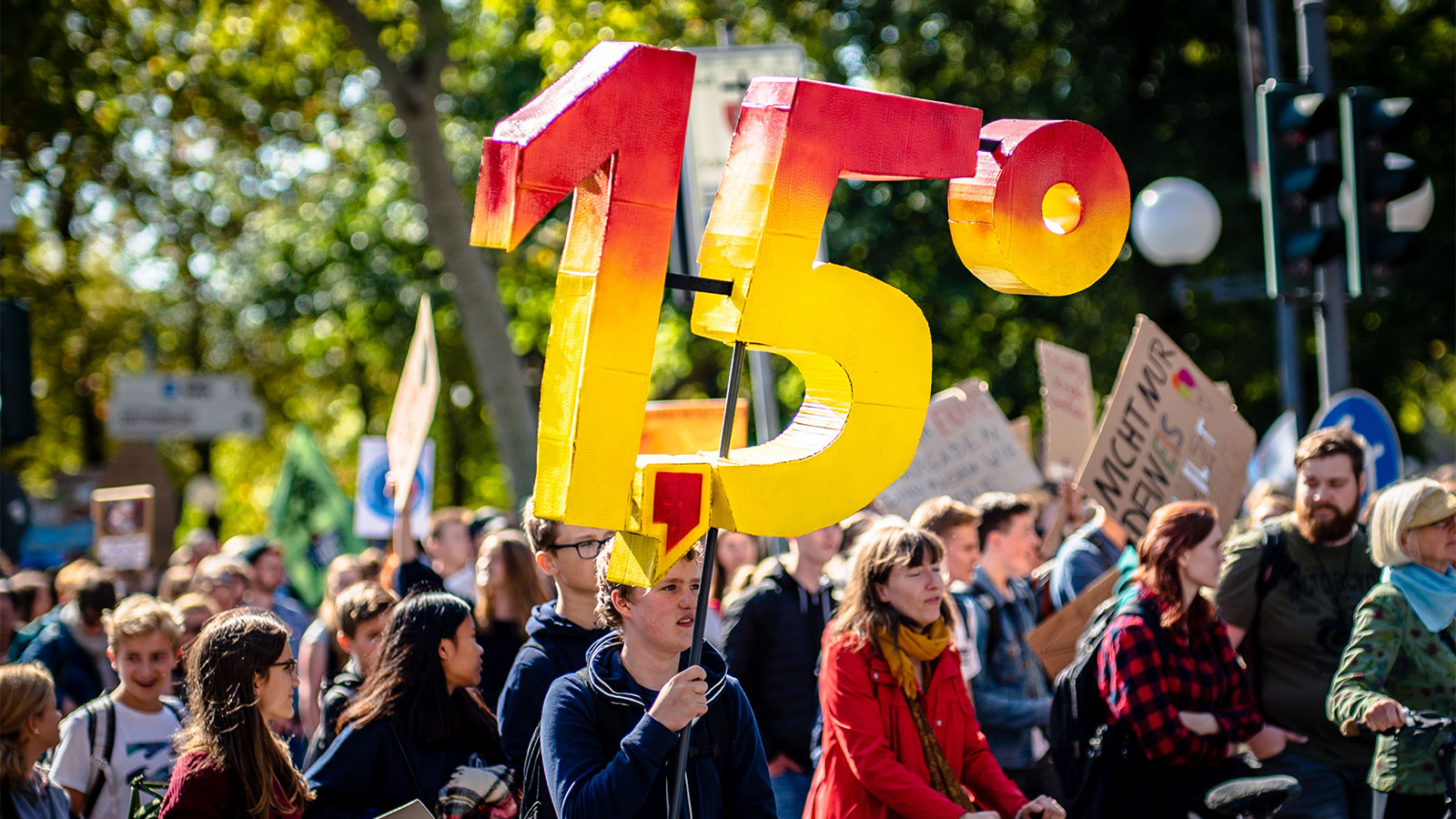 large group of protesters, central person is holding up a large red and yellow 3-D sculpture of the number 1,5º