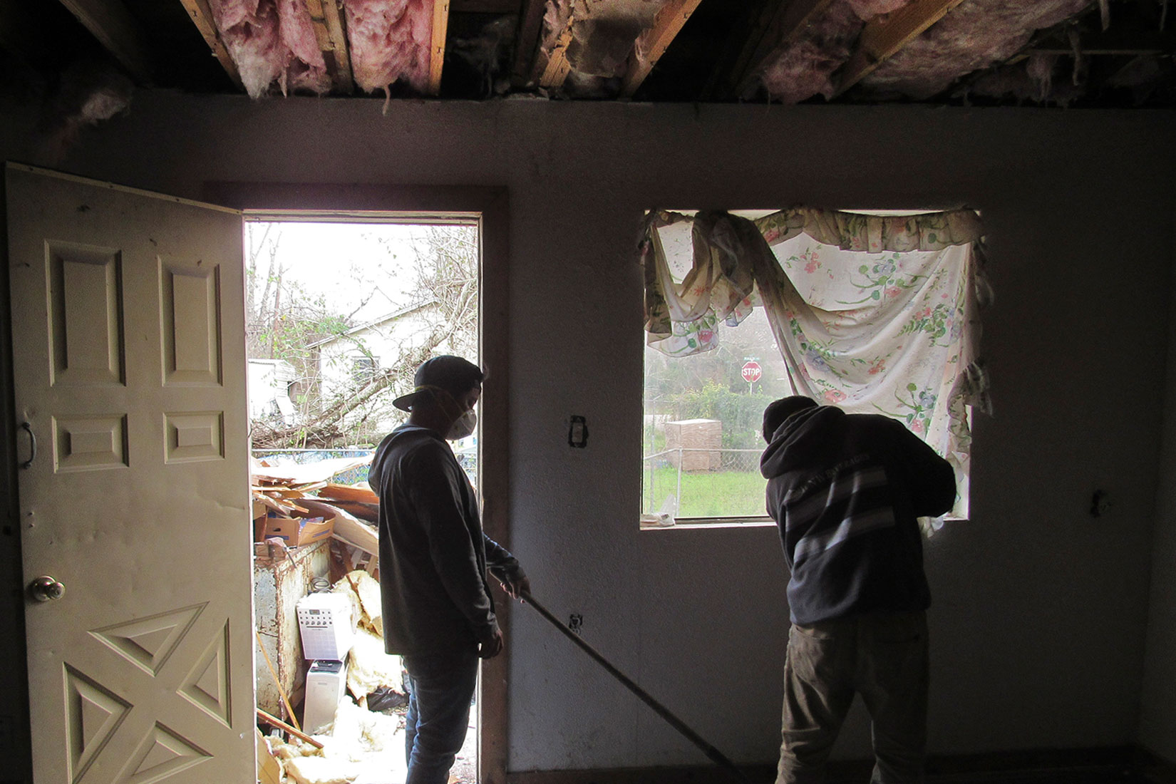 Two people repairing the interior of a home damaged by Hurricane Harvey; a pile of ruined belongings sits outside the doorway and the ceiling is exposed showing insulation