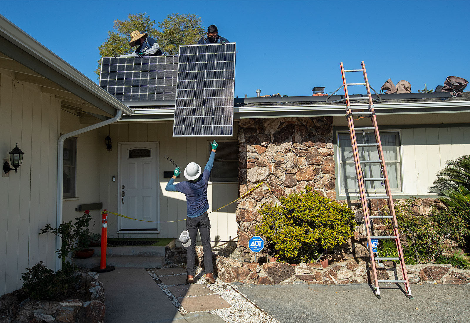 Two men on the roof of a home installing solar panels, one man on ground lifting solar panel to roof