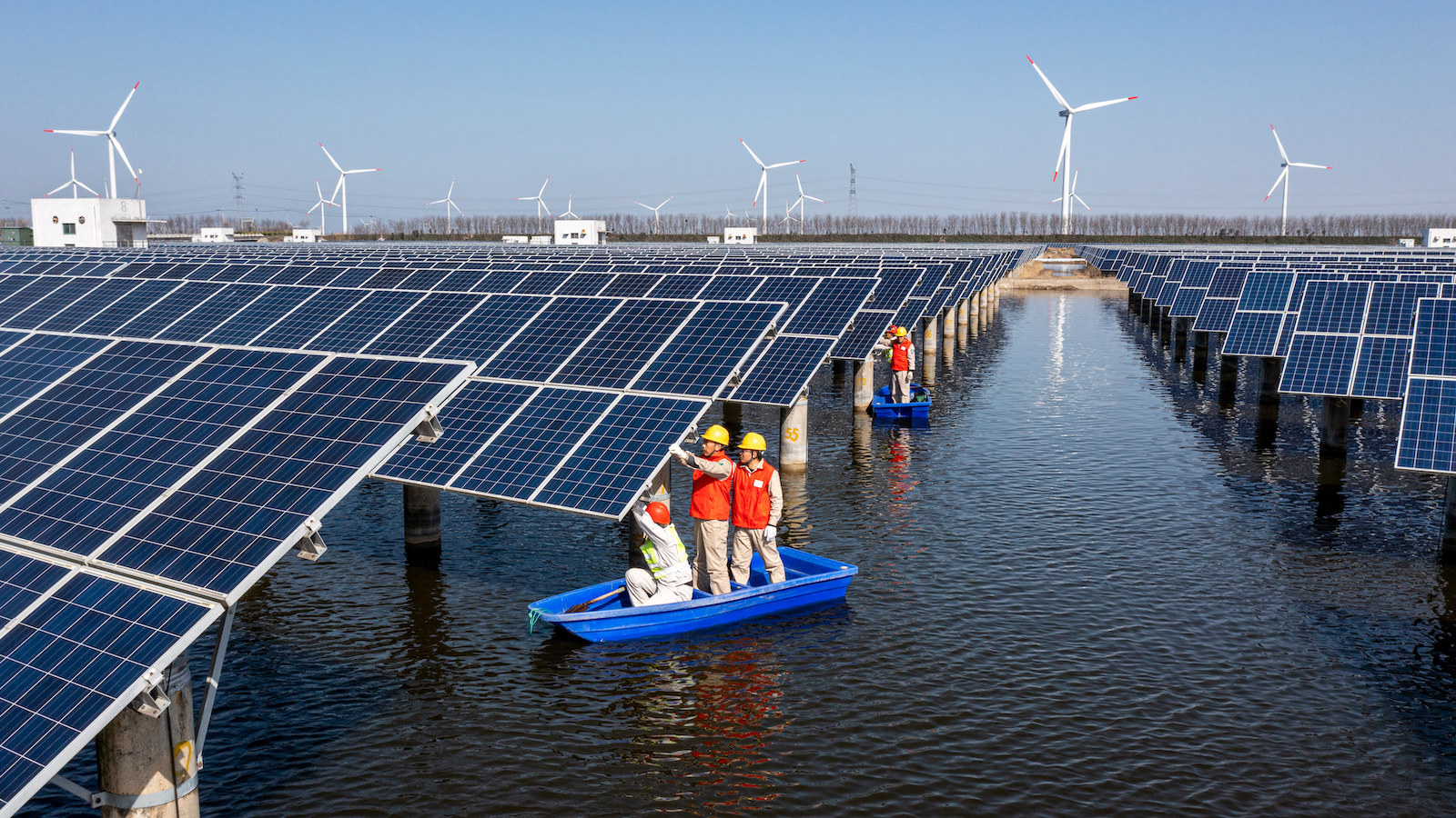 A crew on a boat inspects solar panels with wind turbines in the background