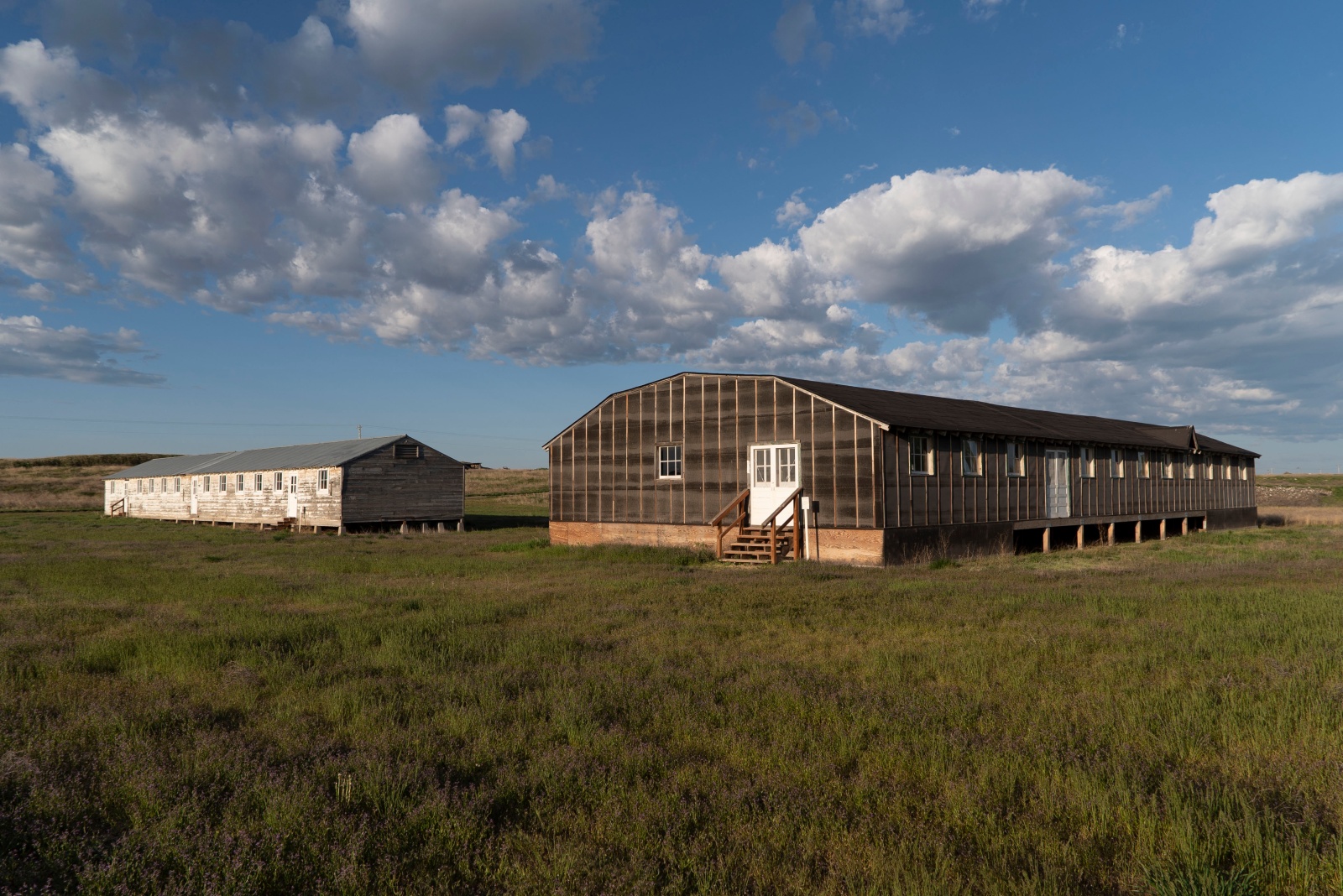 Two low, long buildings in an open grassy plain. There are clouds and a wide horizon surrounding the buildings.