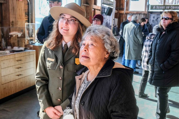 A photo of two women stand and look at something together. The younger woman, left, wears an NPS ranger uniform. An elderly, Japanese American woman stands beside her.