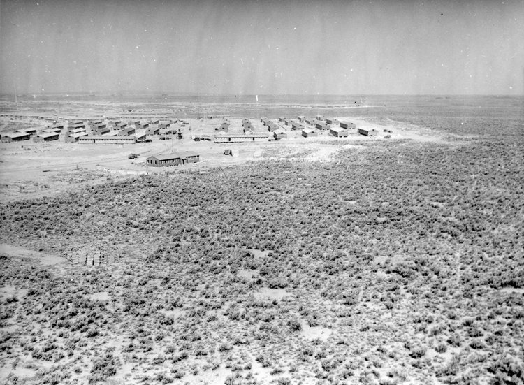A black-and-white photo of an array of buildings surrounded by wide, empty desert and sagebrush.
