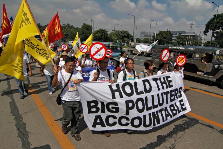 Six people march while holding a sign that says "Hold the big polluters accountable" in capital letters. They lead a line of protesters carrying flags with signs protesting major oil and gas companies.