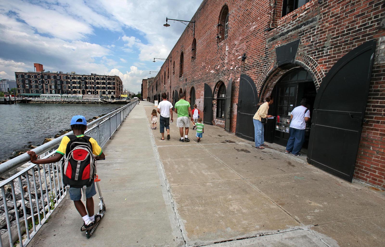 a boy on a scooter rides on a concrete path with water on the left and a red brick building on the right
