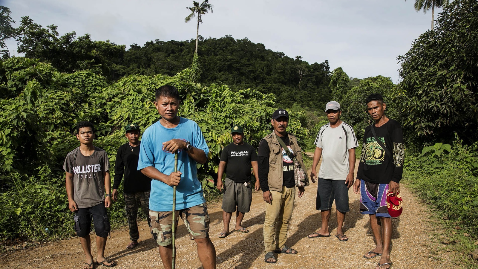 A group of people stand defensively in front of a tropical forest.