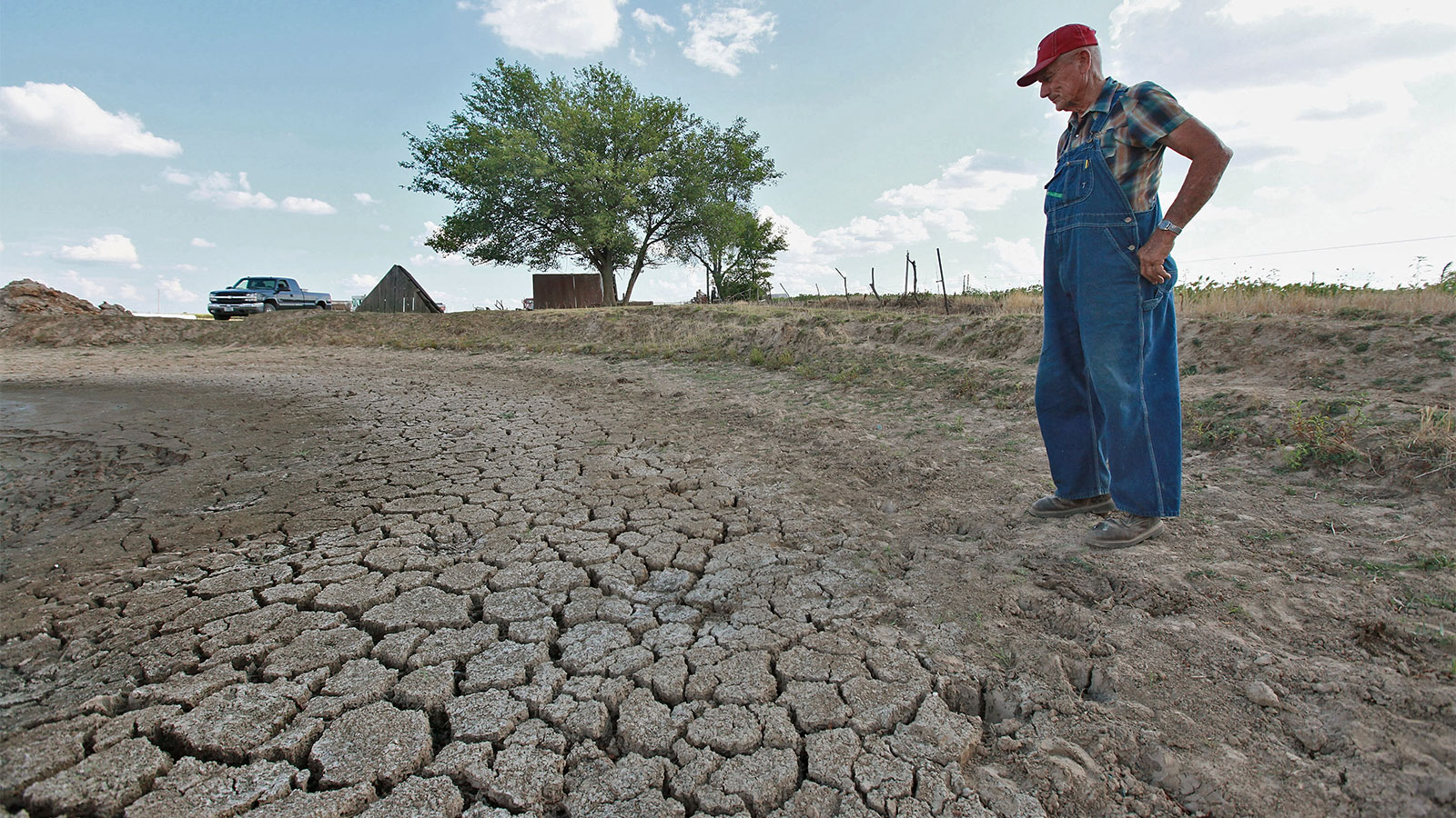 A male farmer wearing overalls and a red cap looking down at dry, cracked earth caused by severe drought