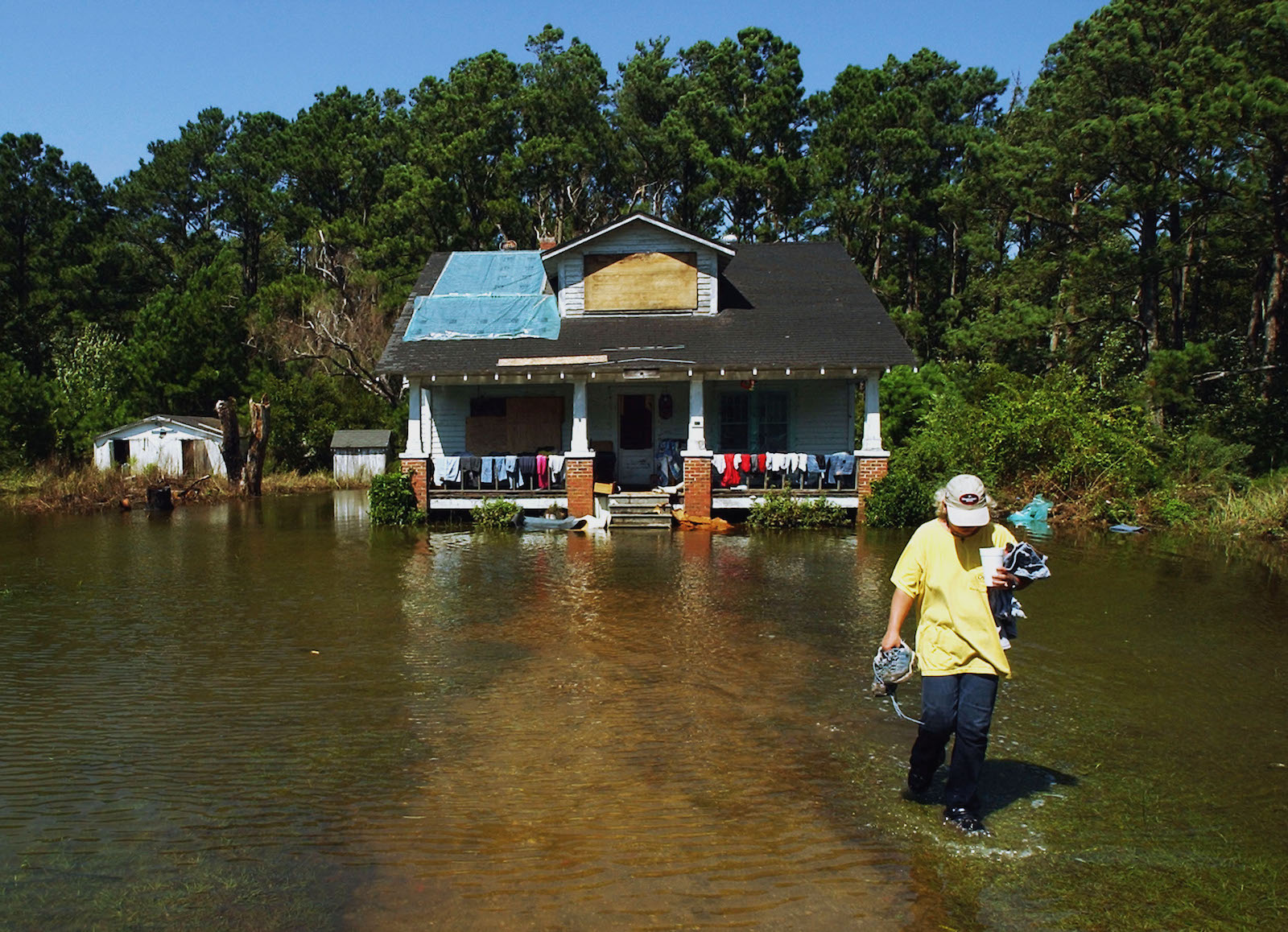 a woman in a hat and t-shirt walks through flood water in front of a patched up house