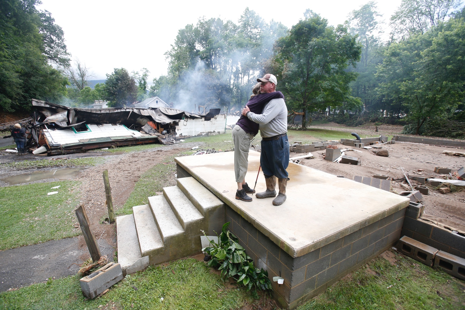 Two people hug on a front stoop, the house behind it demolished, with another collapsed house in the background