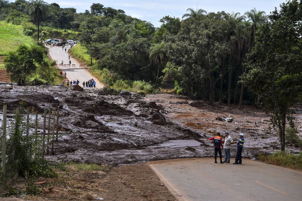 A hilly road surrounded by lush trees is covered with a thick flow of mud. People look onto the damage.