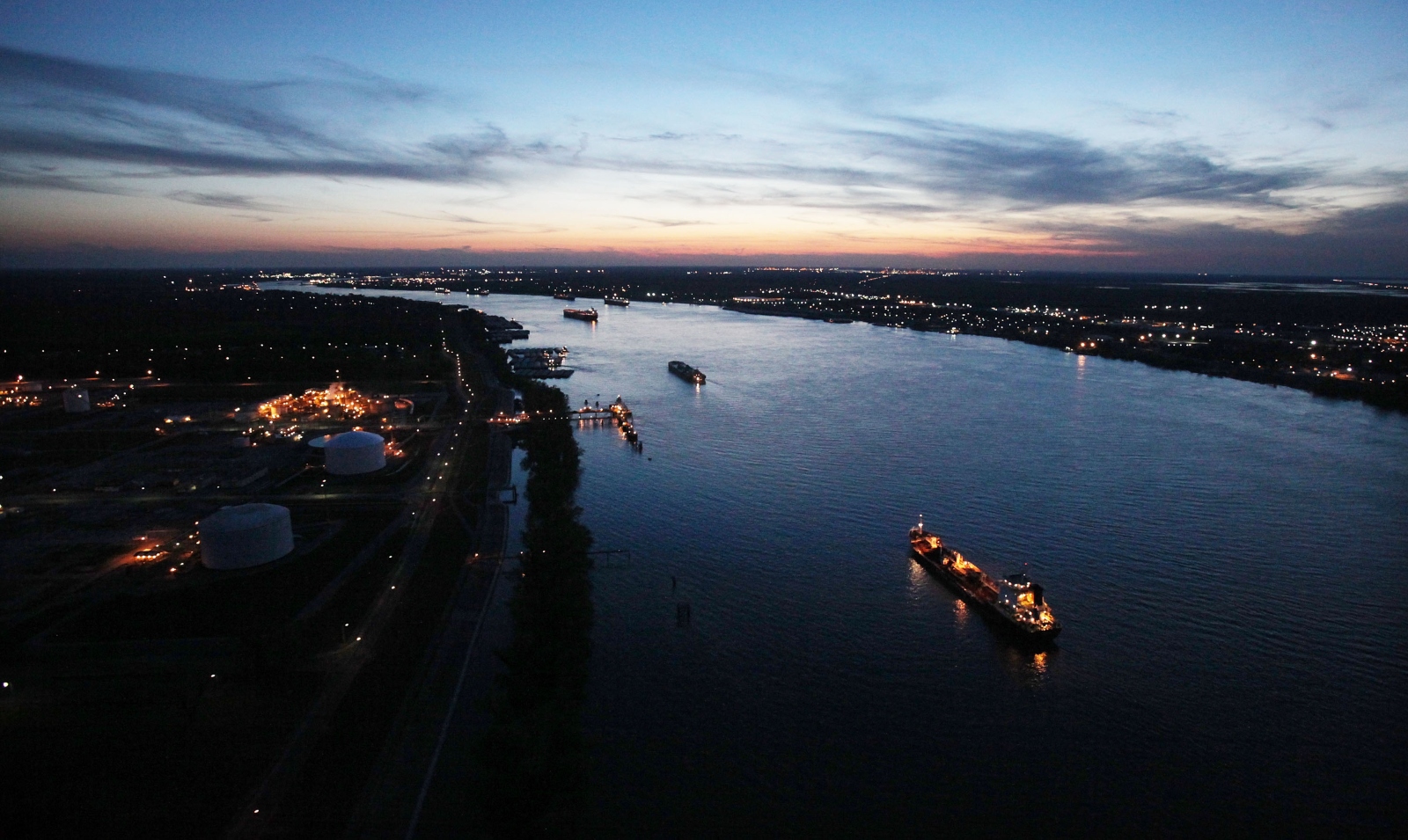 A wide photo of the Mississippi River from above, at twilight. Several cargo ships are in the river.