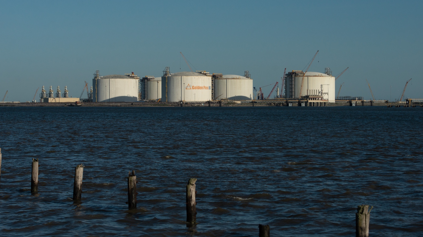 A row of low, wide cylindrical storage tanks against dark water with an evening sky in the background