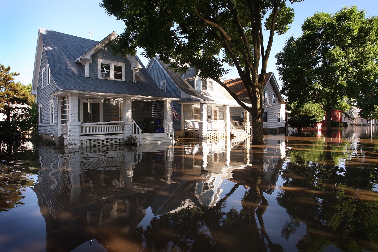 A row of homes on a tree-lined residential street with flood water up to the porches