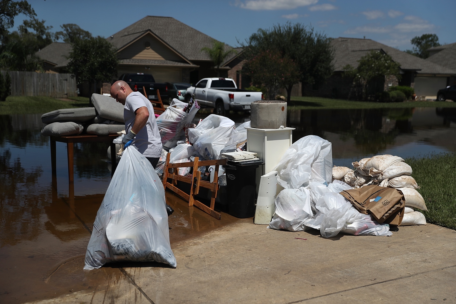 Man pulls garbage bags full of water-damaged household items down sunny driveway against flooded yards.