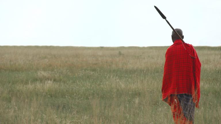 Maasai person with spear and blanket standing in tall grass