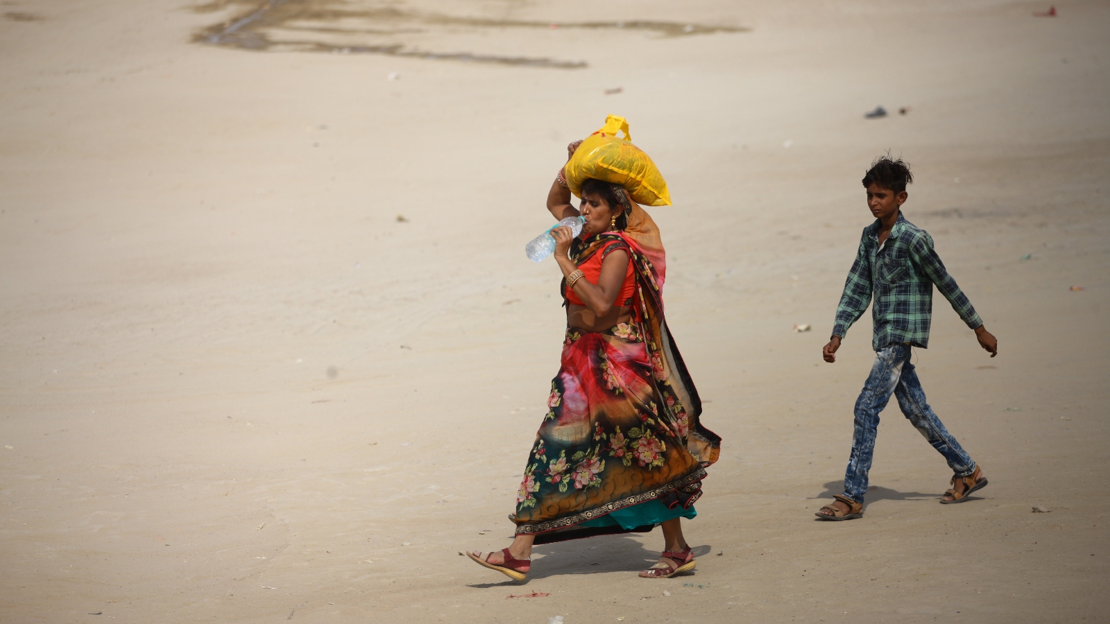 Two people walk in the sand, drinking water and wearing layers to protect from the sun.